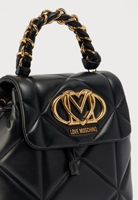 Μαύρη δερμάτινη τσάντα χειρός με καπιτονέ σχέδιο, που διαθέτει χρυσό λογότυπο και χειρολαβή με αλυσίδα. Ομαλή υφή και δομημένο σχήμα.