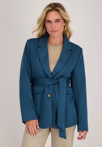 Teal blazer met een strikceintuur, reverskraag en twee voorzakken. De gladde textuur en een enkele gouden knoopdetails verhogen het ontwerp.