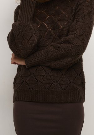 Brun strikket sweater med diamant- og blonddetaljer, båret med matchende brun nederdel, person, der hviler hagen på hånden.