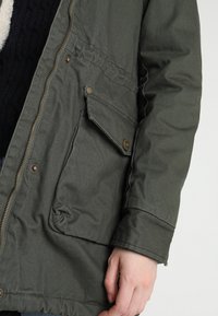 Olivově zelená bunda z lehké látky; má přední zip, druky a boční kapsu s texturovaným povrchem.