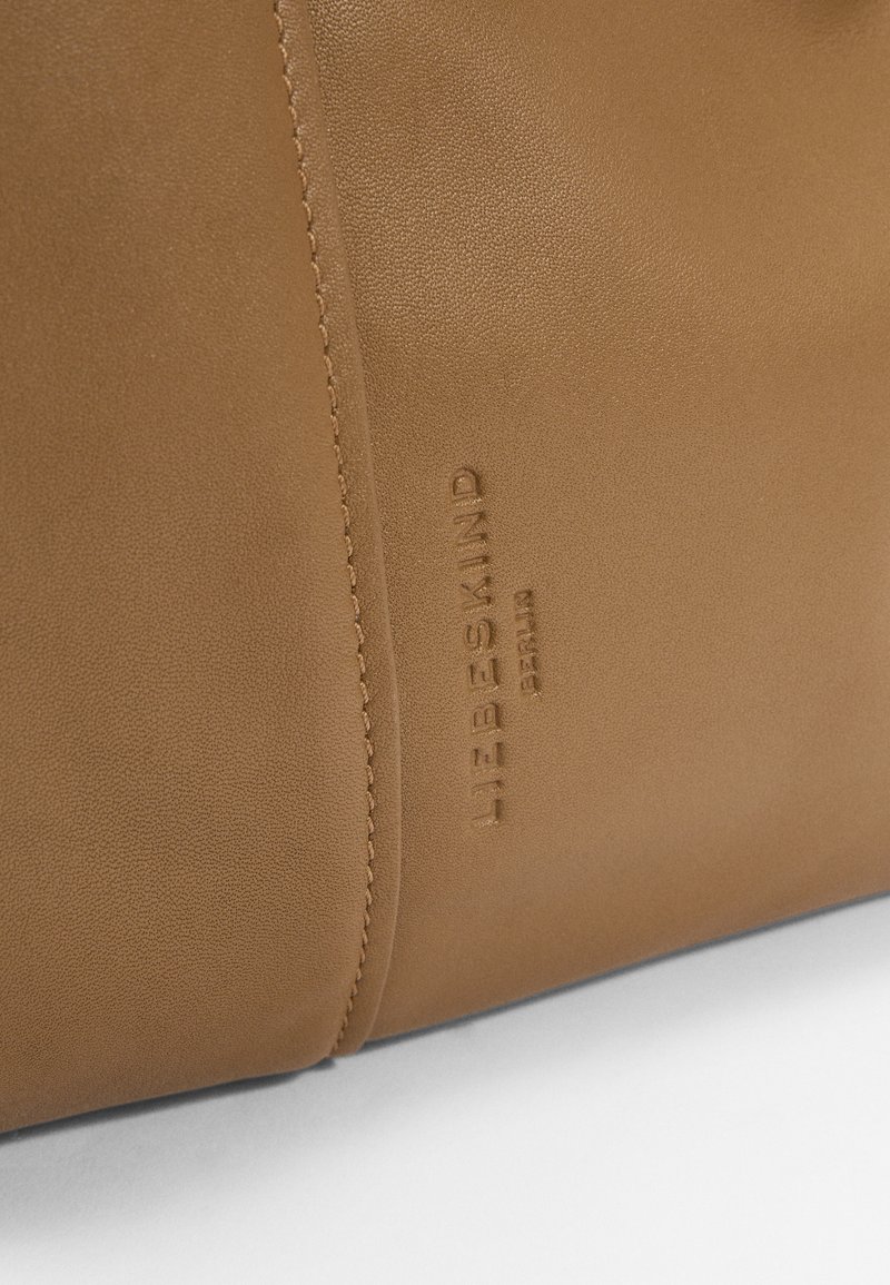 Tasche aus braunem Leder mit glatter Textur, geprägtem Logo und kontrastierenden Nähten. Verfügt über ein minimalistisches Design und eine weiche Form.