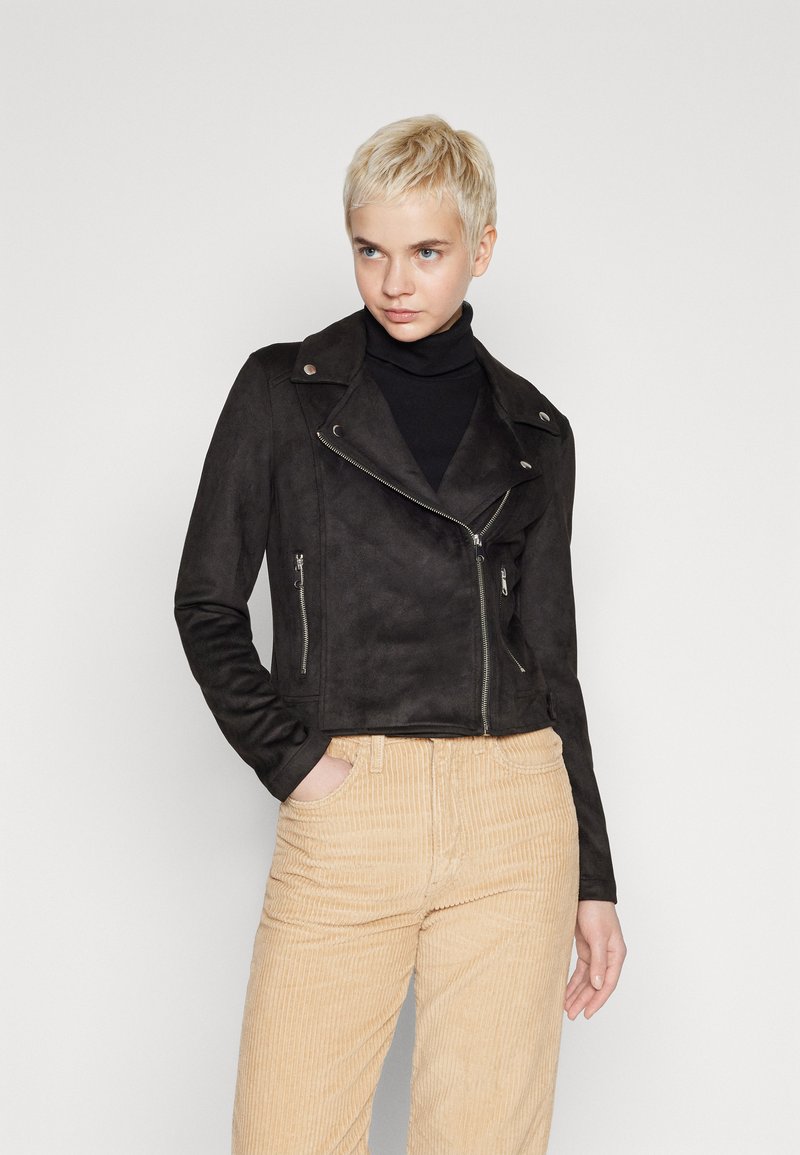 Zwarte suède motorjack met asymmetrische rits, drukknoopkraag en twee zijzakken, gecombineerd met beige corduroy broek.