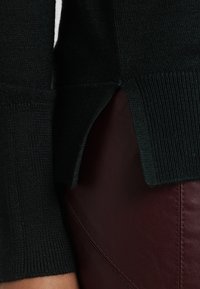 Schwarzer Strickpullover mit strukturierter Optik, seitlichen Schlitzdetails und geripptem Saum, kombiniert mit einem burgunderfarbenen Lederoberteil, das kontrastierende Farben zeigt.