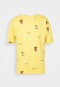 Żółty bawełniany t-shirt z rozproszonymi logo Nike oraz napisem "JUST DO IT" w kolorze bordowym i ciemnopomarańczowym. Okrągły dekolt, krótkie rękawy.