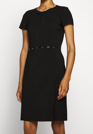 Kvinde iført en tætsiddende, kortærmet, knælængde sort kjole med et diskret dekorativt bælte i taljen mod en ensfarvet baggrund.