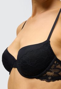 Černá krajková podprsenka s vycpávanými košíčky, zdobená propracovanými krajkovými detaily podél okrajů a stříbrným akcentem na bočním popruhu.