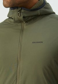 Veste zippée vert olive avec col haut, fabriquée en tissu léger. Présente un logo et une fermeture éclair contrastante à l'avant.