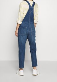 Jeansoveraller med vida ben, utrustade med två bakfickor, justerbara axelband och en blekt blå färg, kombinerade med en randig gul och vit skjorta.