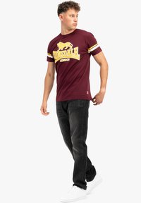 Rudai marškinėliai su geltonu tekstu ir liūto grafika, trumpomis rankovėmis su dryžuotais akcentais. Dėvimi su juodais džinsais ir baltais sportbačiais.