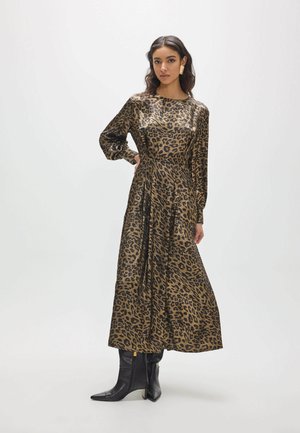 Femme portant une longue robe marron et noire à imprimé léopard avec des manches longues et des bottes noires pointues sur un fond uni.