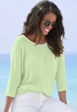 Hellgrünes, locker sitzendes Oberteil mit Dreiviertelärmeln und Rundhalsausschnitt. Glatte Textur, kombiniert mit weißen Hosen und Sonnenbrille.