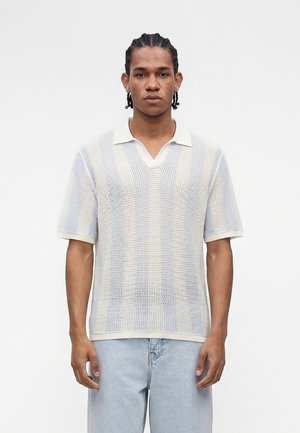 Junger Mann mit geflochtenem Haar, der ein weißes kurzärmeliges Mesh-Poloshirt und hellblaue Jeans trägt, steht vor einem schlichten Hintergrund.
