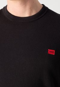 Gros plan d'une personne portant un pull noir à col rond avec une petite étiquette rectangulaire rouge « HUGO » sur la poitrine.