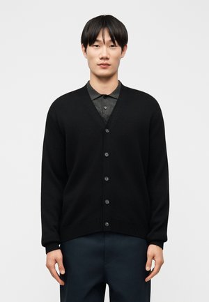 Nuori mies seisoo musta napillinen neuletakki päällään, jonka alla on tummanharmaa kauluksellinen paita ja tummat housut, yksiväristä taustaa vasten.