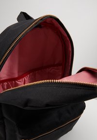 Zwarte rugzak met een gouden rits, voorzien van een rood gestreepte binnenvoering en een binnenzak. De stof is stevig en heeft een structuur.