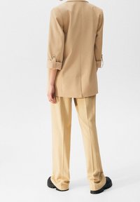 Personne portant un pantalon beige sur mesure et un blazer assorti aux manches retroussées, debout devant un fond blanc uni.