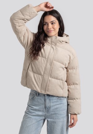 Frau mit langen dunklen Haaren, die eine beige Daunenjacke und hellblaue Jeans trägt, hebt einen Arm über dem Kopf und lächelt leicht.