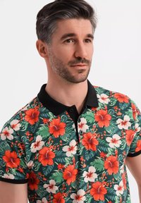Kortærmet polo skjorte med et levende blomsterdesign med røde og hvide blomster på en grøn baggrund, sort krave og knaplukning.