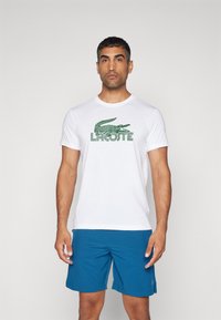 Camiseta blanca de manga corta con el logo del cocodrilo verde de Lacoste y texto en la parte frontal, combinada con pantalones cortos azules. Tejido suave, diseño ajustado.