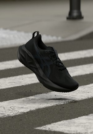 Schwarzer Sportschuh mit einer strukturierten Oberseite, auffälligem Logo und gedämpfter Sohle, schwebt über einem gestreiften Zebrastreifen.