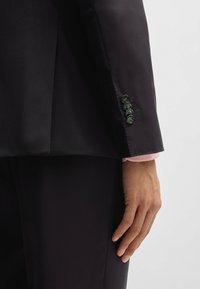 Zwarte blazer van een gladde, glanzende stof. Bevat een roze manchet die eronder zichtbaar is. Vijf donkere knopen op de manchet, die het op maat gemaakte ontwerp benadrukken.