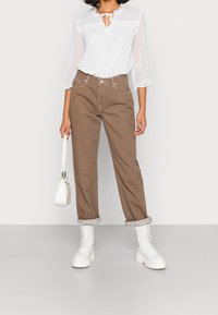White, textured long-sleeve blouse with tie neckline; brown high-waisted trousers; white chunky lace-up boots; small white handbag.