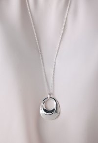 Silver pendant necklace with rounded, abstract open circle design on a fine chain against a smooth light fabric background.