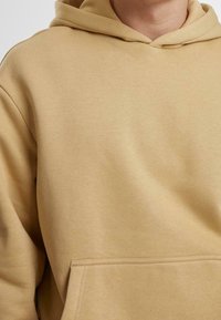 Detailní záběr na osobu ve béžové mikině s kapucí, s viditelným prošíváním kapuce a přední kapsy.