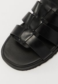 Black sandal featuring multiple wide, overlapping straps made of smooth material. Flat sole with a textured surface and rounded edges.