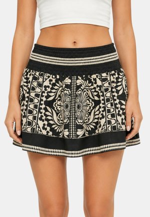Donna che indossa un crop top bianco e una minigonna nera con motivi floreali e geometrici beige, in piedi con le braccia rilassate lungo i fianchi.