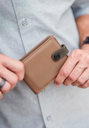 Braune Stoffgeldbörse mit strukturierter Oberfläche, Reißverschluss und grünem Akzentriemen, in einer Hand gehalten vor einem hellblauen Hemd.