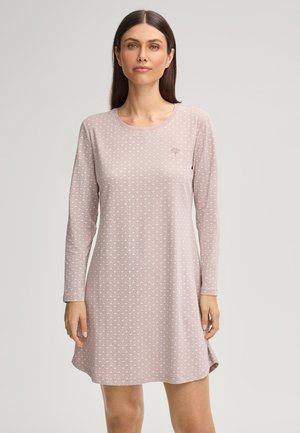 Vrouw met steil donker haar, die een lichtroze jurk met lange mouwen en patroon draagt, staand tegen een effen lichte achtergrond.