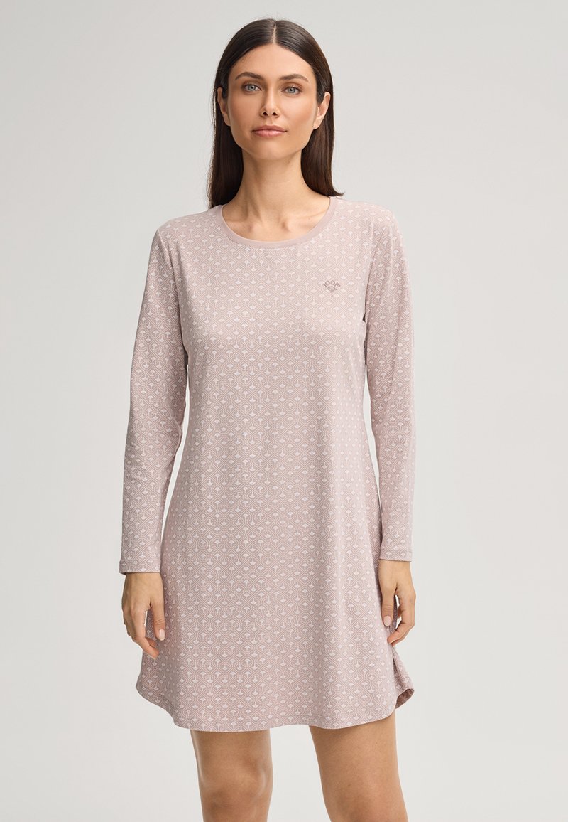 Femme aux cheveux bruns raides portant une robe à manches longues rose clair à motifs, debout devant un fond clair uni.