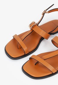 Sandali in pelle color cuoio con cinturini sottili, punta quadrata e cinturino alla caviglia con fibbia. La suola nera mette in risalto il design.