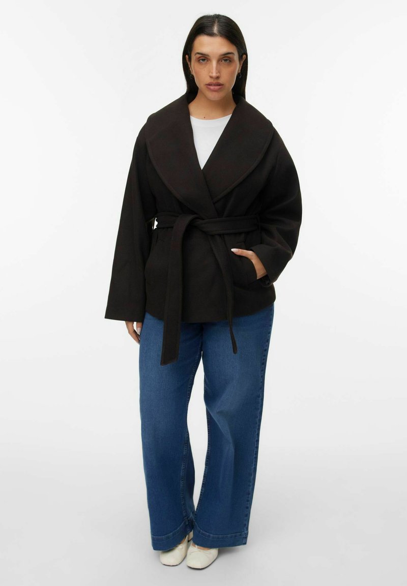 Abrigo negro con un amplio cuello, cintura ceñida por un cinturón y mangas largas, combinado con unos vaqueros azul de pierna ancha y calzado blanco.