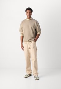 Béžové oversized tričko, khaki kalhoty a tenisky se zelenými akcenty. Hladká tkanina, volný střih a žebrované manžety na rukávech.