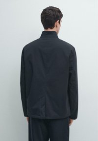 Sort, let, hofte-lang jakke med høj krave, lige snit og minimalistisk design. Har en slids bagpå for nem bevægelse.