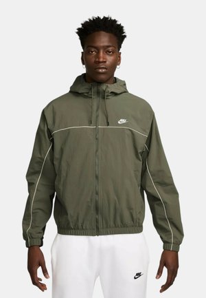 Hombre con chaqueta con capucha Nike verde oliva con ribetes blancos y pantalones deportivos Nike blancos, de pie con los brazos relajados a los lados.