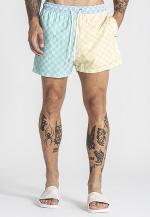 Mand med tatoverede ben iført pastelfarvede blå og gule ternede shorts og hvide slidesandaler, stående foran en ensfarvet baggrund.