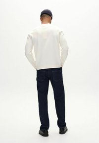Camisa blanca de manga larga con cuello redondo, combinada con jeans de mezclilla azul oscuro y zapatos negros, vista desde la parte de atrás.