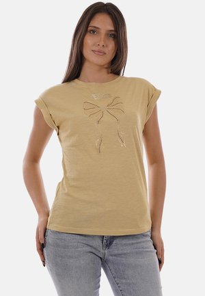 Donna che indossa una t-shirt beige a maniche corte con design a fiocco di strass e scritta "YES ZEE", abbinata a jeans grigio chiaro, in piedi davanti a uno sfondo bianco.