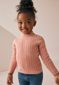 Baby-pink gestrickter Pullover mit hohem Ausschnitt, der über vertikale Rippungen und Zopfmuster verfügt, kombiniert mit blauen Jeans.