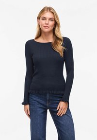 Femme aux longs cheveux blonds portant un pull côtelé bleu foncé ajusté avec des bords à volants et un jean bleu, debout devant un fond blanc.