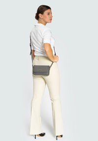 Borsa a tracolla in pelle grigia con superficie texturizzata, dotata di chiusura a pattina e tracolla regolabile, indossata da una persona in pantaloni fusciacca beige.