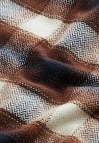 Nærbillede af blødt uldstof med et brunt, cremefarvet, sort og hvidt herringbone-rutet mønster og synlig tekstur.