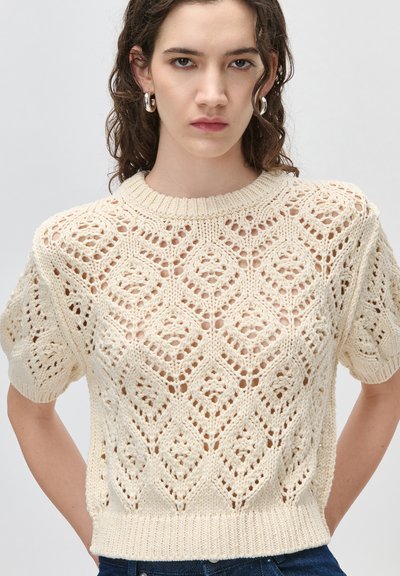 Jeune femme aux cheveux bouclés et foncés portant des boucles d'oreilles argentées en forme de cerceau et un pull court à manches courtes en crochet de couleur crème avec des motifs en losange.