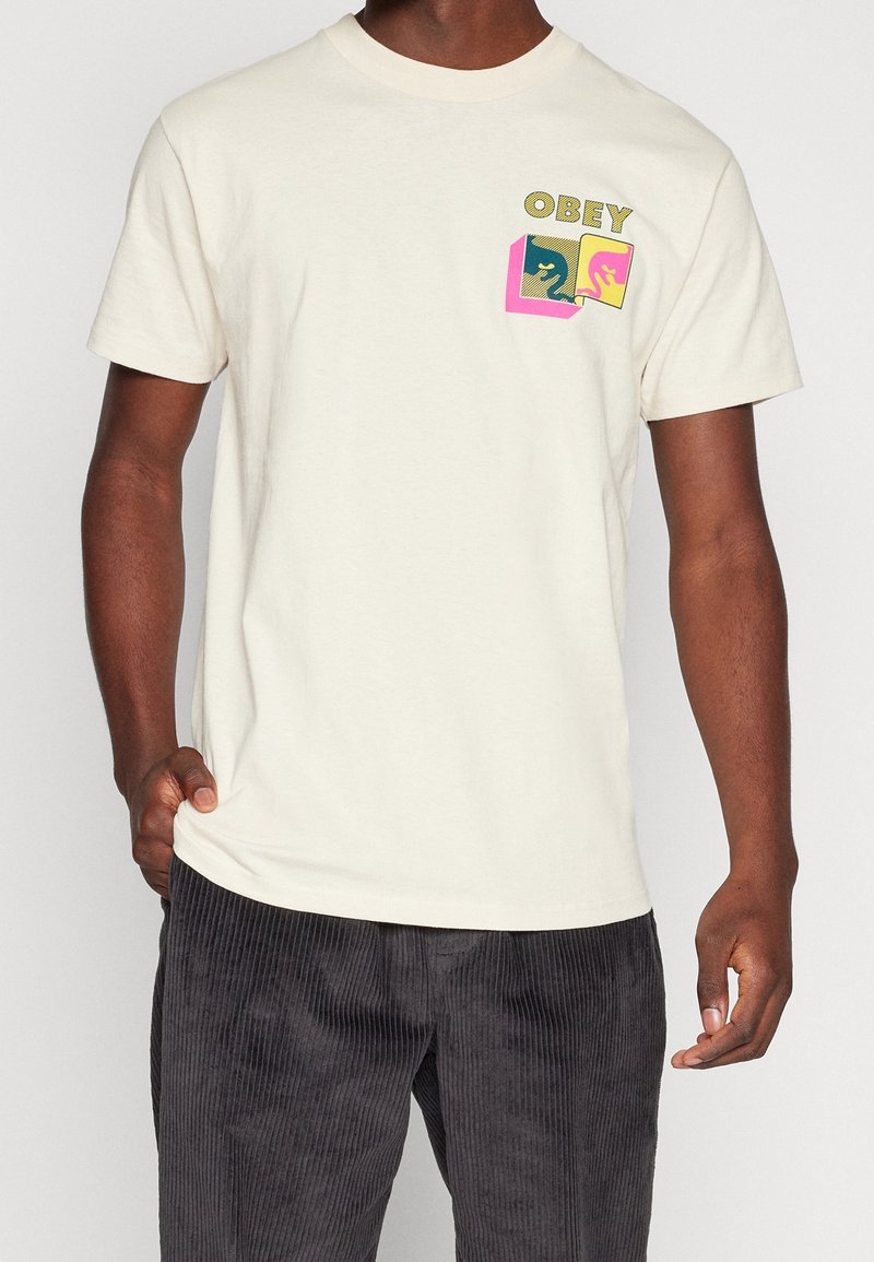Kremowa bawełniana koszulka z krótkim rękawem, z wielokolorowym nadrukiem "OBEY" na lewej stronie w okolicy kieszonki. Luźny krój.