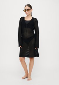 Black mesh cover-up dress with long sleeves, tie-front detail, and semi-transparent texture, paired with square-shaped, tinted sunglasses.