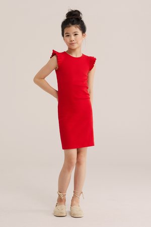 Fată stând cu mâna pe șold purtând o rochie roșie fără mâneci cu umeri plisati și pantofi espadrile bej cu șireturi.