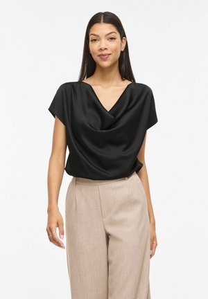 Pitkähiuksinen nainen mustassa satiinissa drapeeratussa paidassa ja korkeavyötäröisissä beigen sävyisissä housuissa valkoista taustaa vasten.